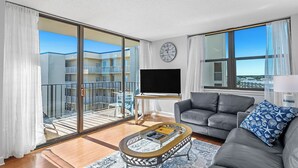 En moderne stue med en grå sofa, et sofabord med magasiner, et fjernsyn på et stativ og en balkon med udsigt til bygninger.