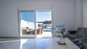 A modern living room with a glass door leading to a pool area, a white sofa, and a vase with flowers on a table. - Vigla Kiotari - B, Villa (Greece)