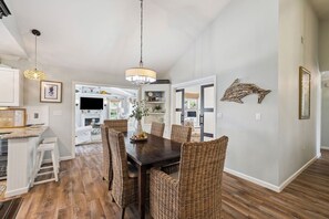 A dining area with a dark wooden table and wicker chairs, a hanging light fixture, and a view into a living room with a fireplace.