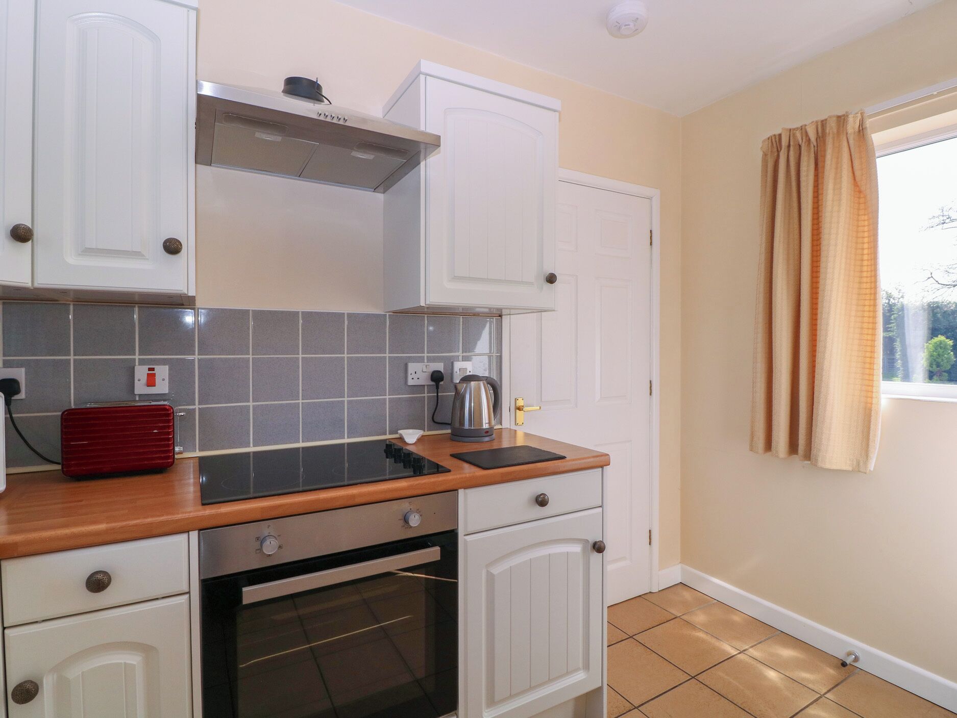 A kitchen with white cabinets, a stove, an oven, a red portable heater, and a window with curtains.