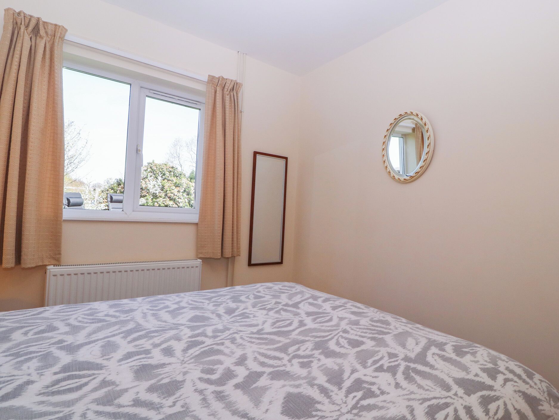 A bedroom with a bed, a window with curtains, a radiator, and a mirror on the wall.