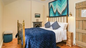 Un dormitorio con una cama, un aparador de madera, una colcha azul acolchada, un otoman azul, una puerta de madera y un cuadro en la pared.