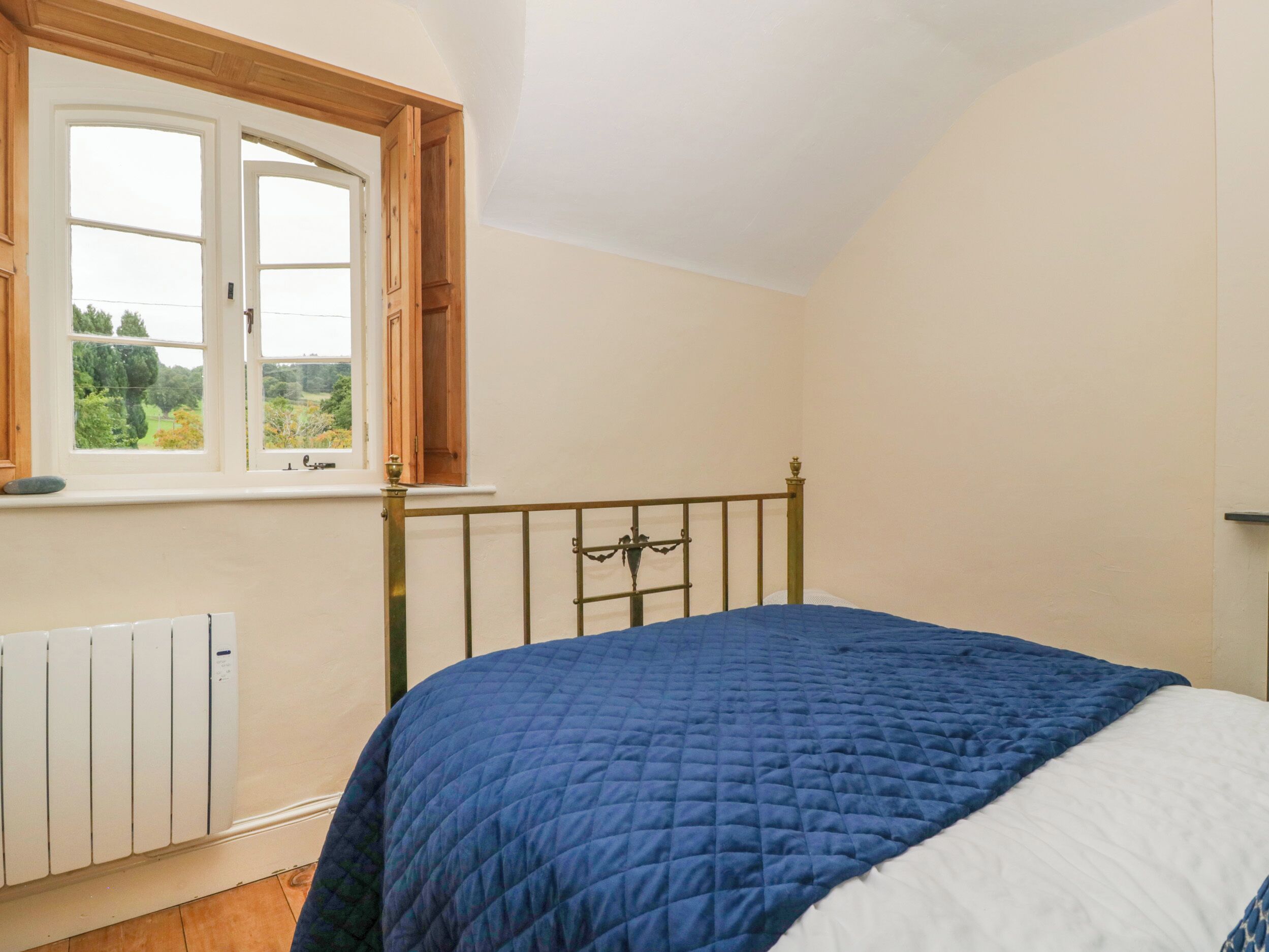 Ein Schlafzimmer mit Bett, Heizkörper, Fenster mit Blick auf Grünflächen und Holzboden.