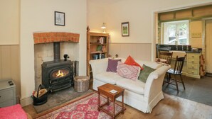Una sala de estar acogedora con chimenea, un sofá blanco con cojines coloridos, una mesa de centro de madera, y una cocina visible a través de una puerta abierta.