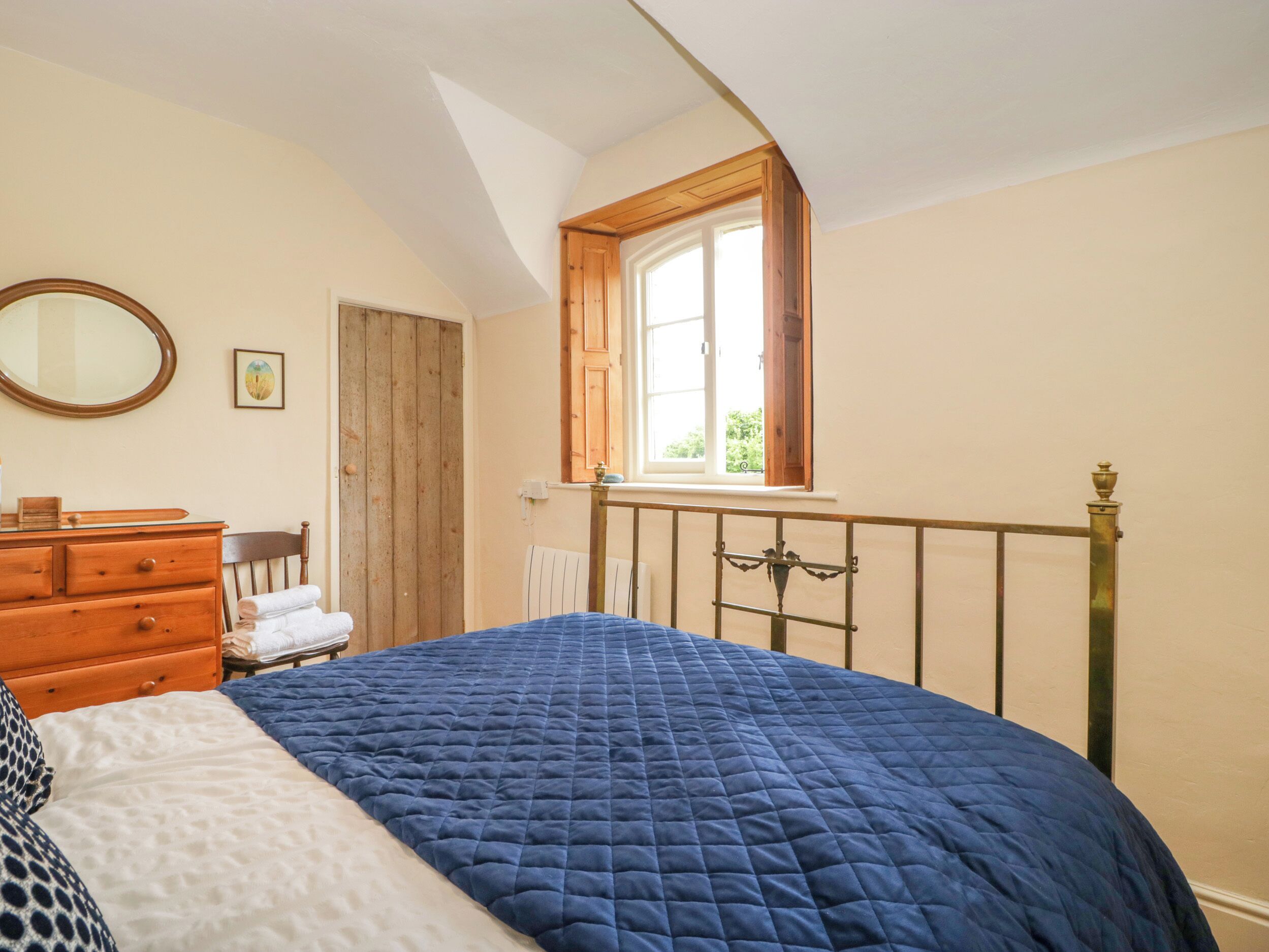 Ein Schlafzimmer mit einem großen Bett, einer Holzkommode, einem Spiegel und einem Fenster mit Blick auf Grünflächen.