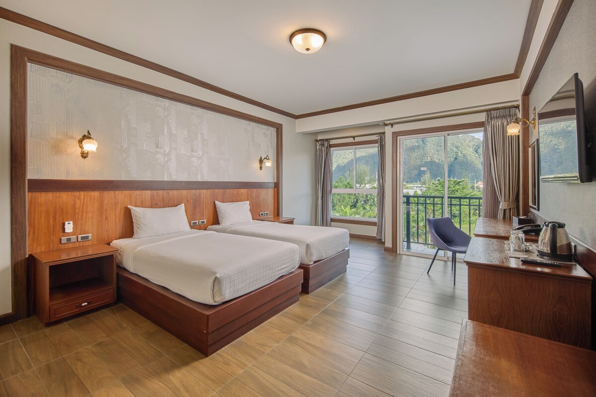 A hotel room with two beds, a desk with a kettle, a chair, and a balcony with a view of greenery.
