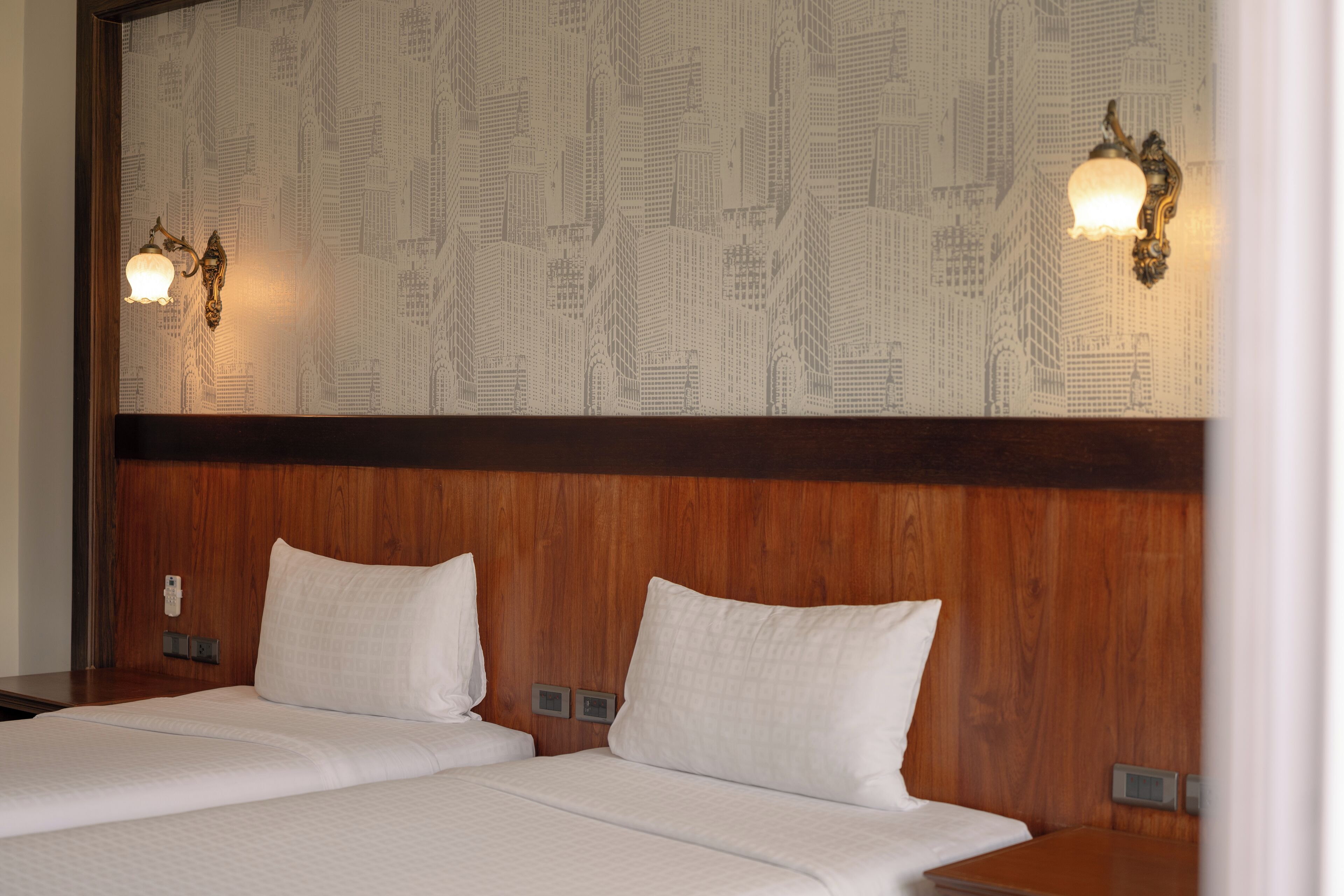 A hotel room with two beds, wooden headboard, patterned wallpaper, and wall-mounted light fixtures.