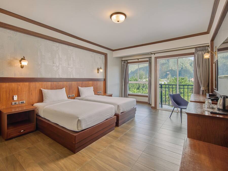 Ein Hotelzimmer mit zwei Betten, einem Schreibtisch mit Wasserkocher, einem Stuhl und einem Balkon mit Blick auf Grünflächen.