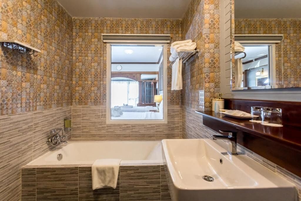 Un baño con una bañera grande, un lavabo y una ventana que da a un dormitorio.