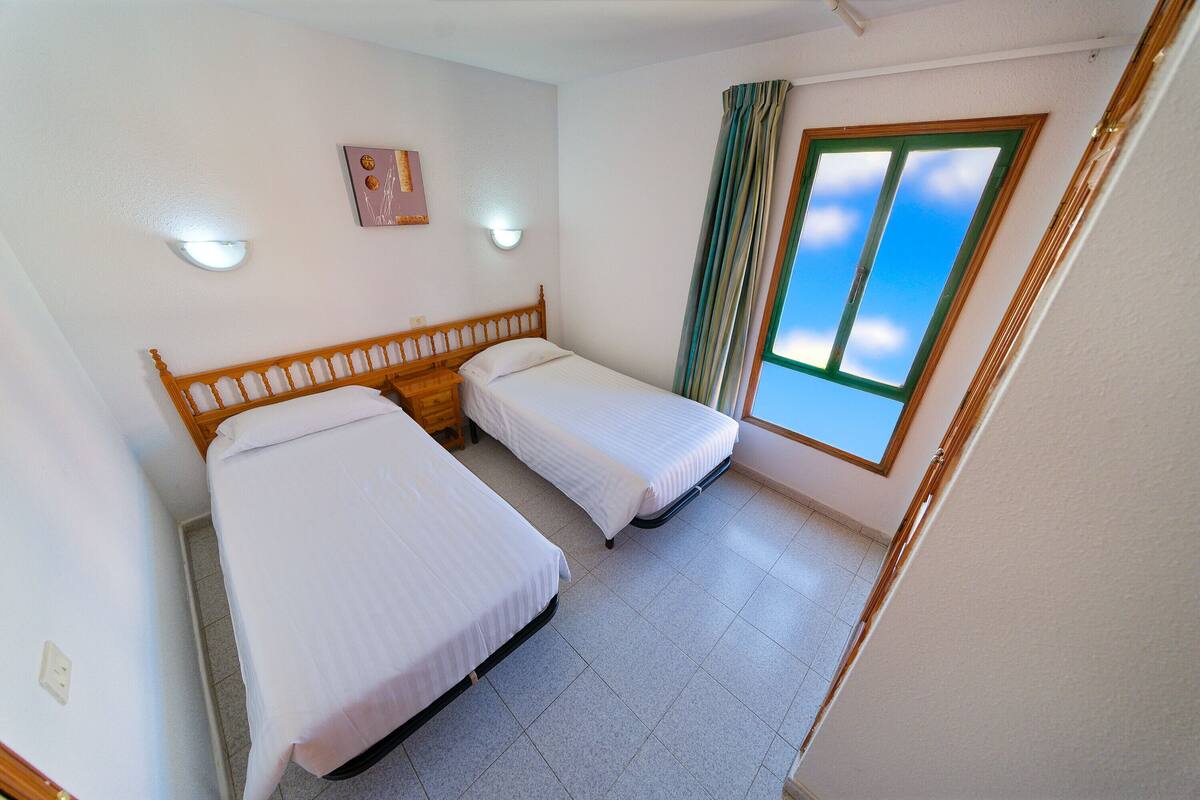 A room with two single beds, a small table, a wall-mounted light, a window with curtains, and a ceiling light.
