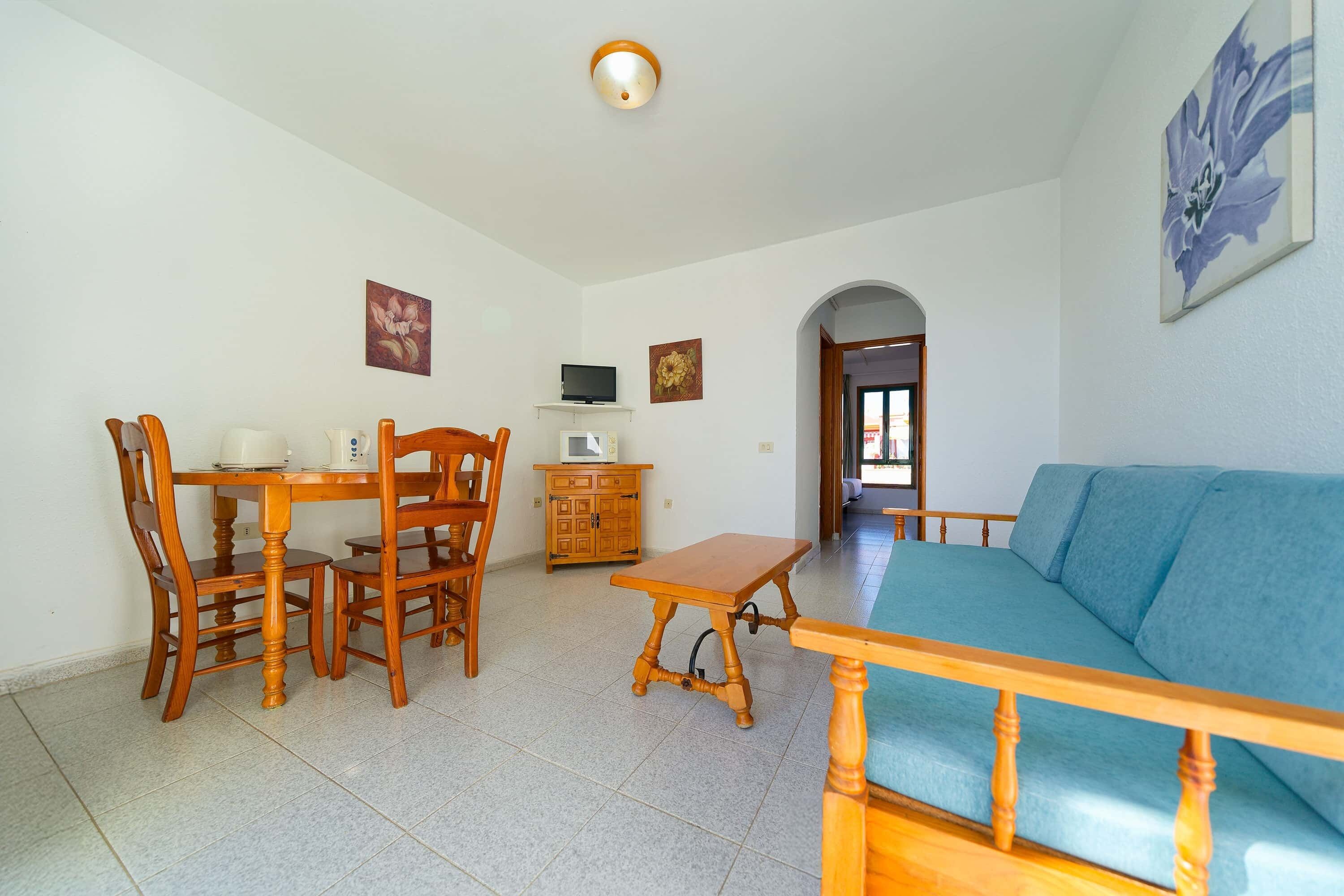 A living room with a blue sofa, wooden dining table with chairs, a small TV on a shelf, and a microwave.