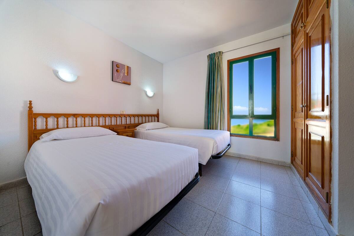A hotel room with two beds, a window with curtains, a wooden wardrobe, and a wall-mounted light fixture.