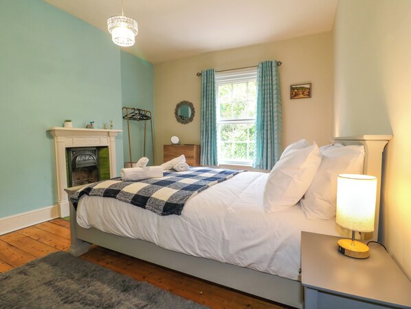 A bedroom with a bed, a nightstand, a lamp, a dresser, a mirror, a window with curtains, a fireplace, and a hanging light fixture.
