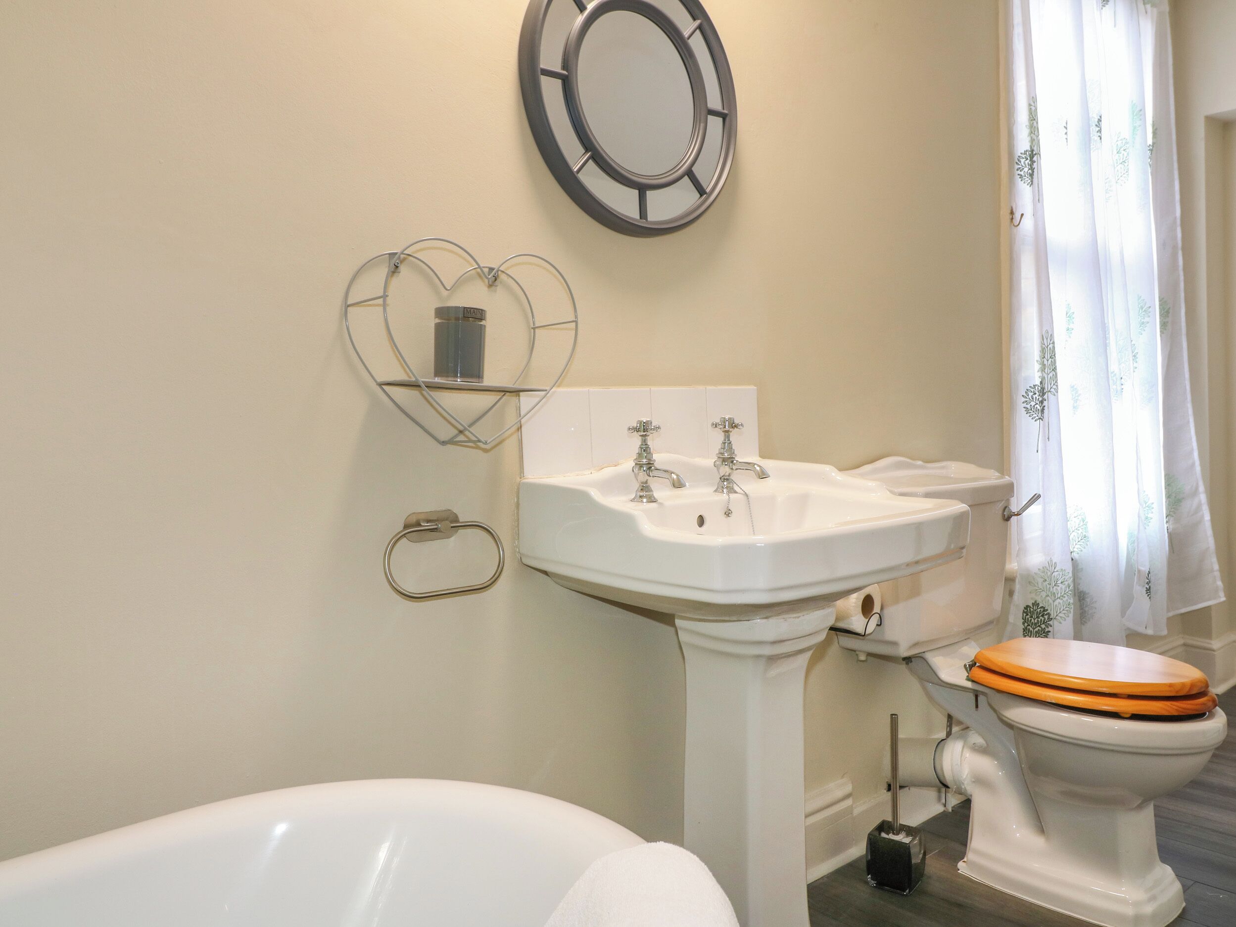 Un baño con una bañera blanca, un lavabo pedestal blanco y un inodoro con asiento de madera. Hay una repisa con forma de corazón sobre el lavabo y un espejo redondo en la pared.