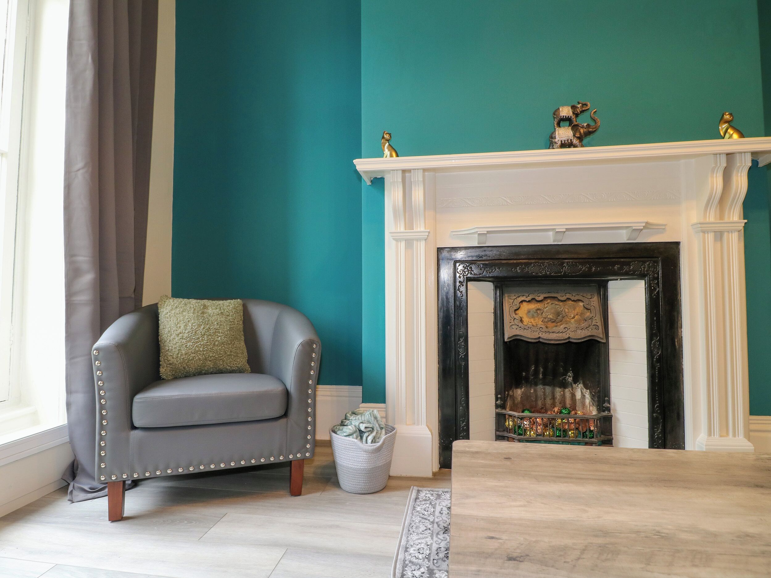 Um quarto com paredes verde-água, uma poltrona cinza com almofada verde, uma lareira branca com objetos decorativos e uma cesta branca no chão.