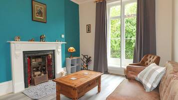 Een woonkamer met een open haard, een bank, een bijzettafeltje en een groot raam met gordijnen.