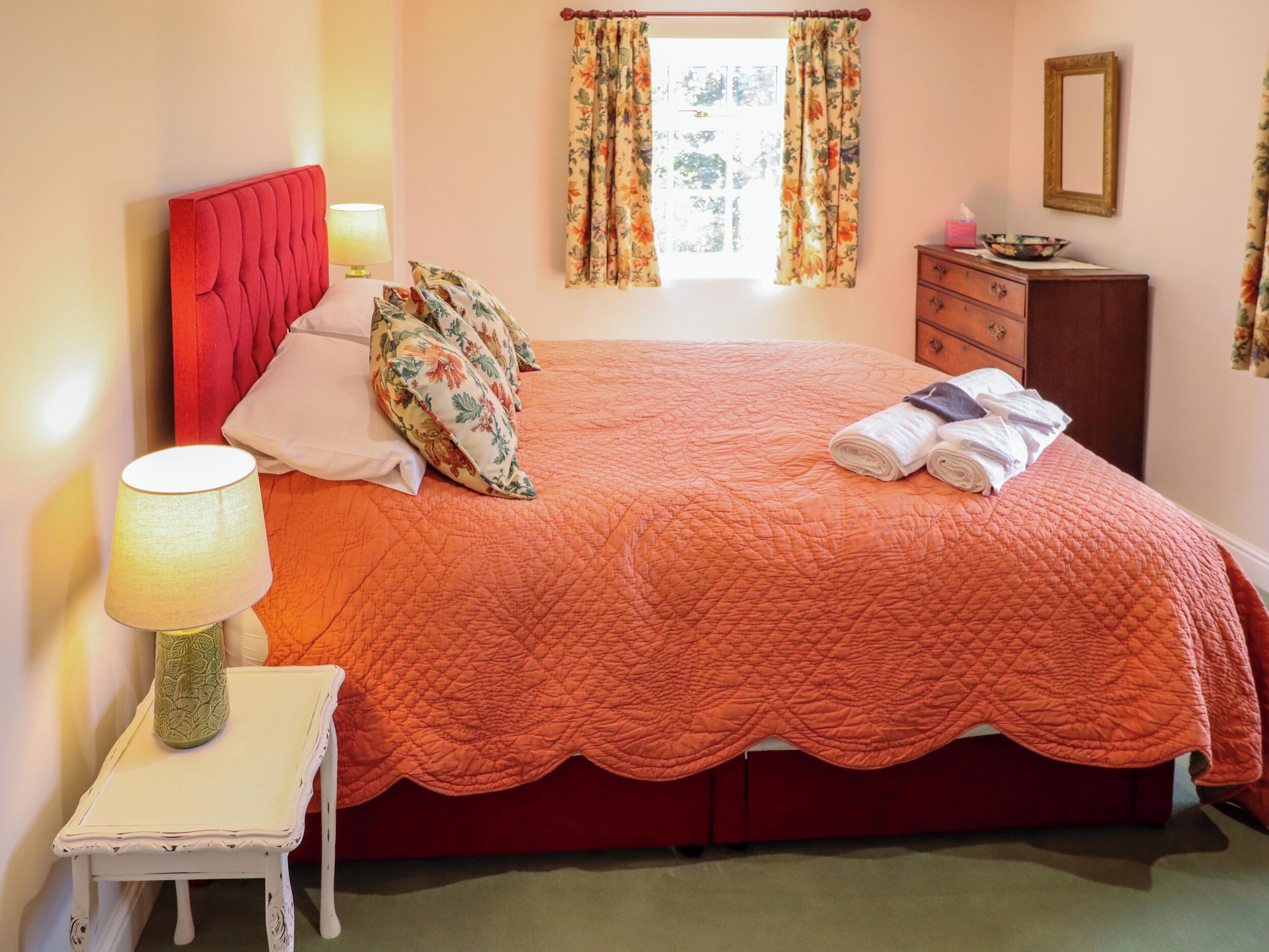 A neatly made bed with a red headboard, a bedside lamp, and floral curtains.