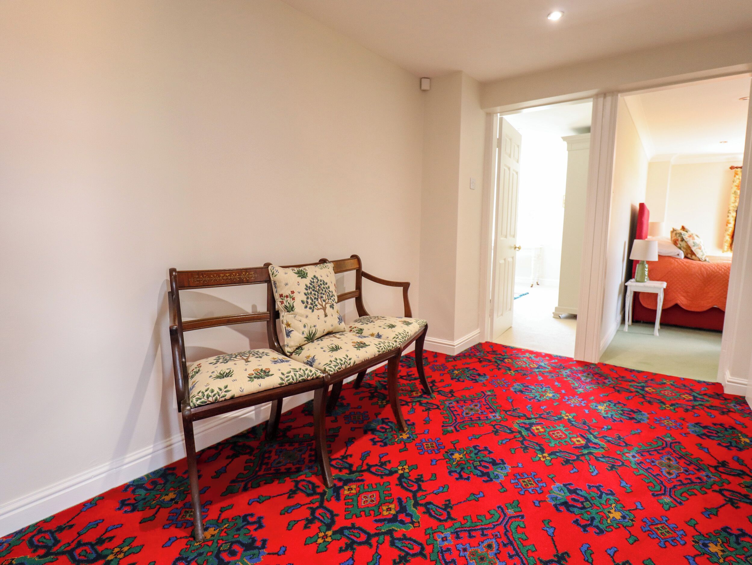 Ein Zimmer mit einem roten Teppich, einer hölzernen Bank mit floralen Kissen und einer weißen Tür, die in einen anderen Raum führt.