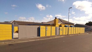 A street with a row of yellow and grey buildings, including a garage and a small structure with a blue roof. - Neo & ruks guest house goodwood (Cape Town)