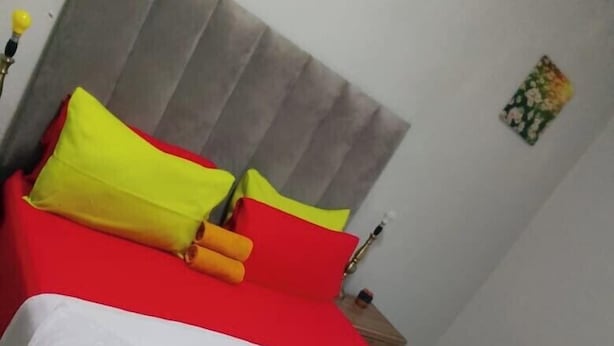 A bed with a red and white bedspread, a grey upholstered headboard, and two pillows, one yellow and one red. - Neo & ruks guest house goodwood (Cape Town)