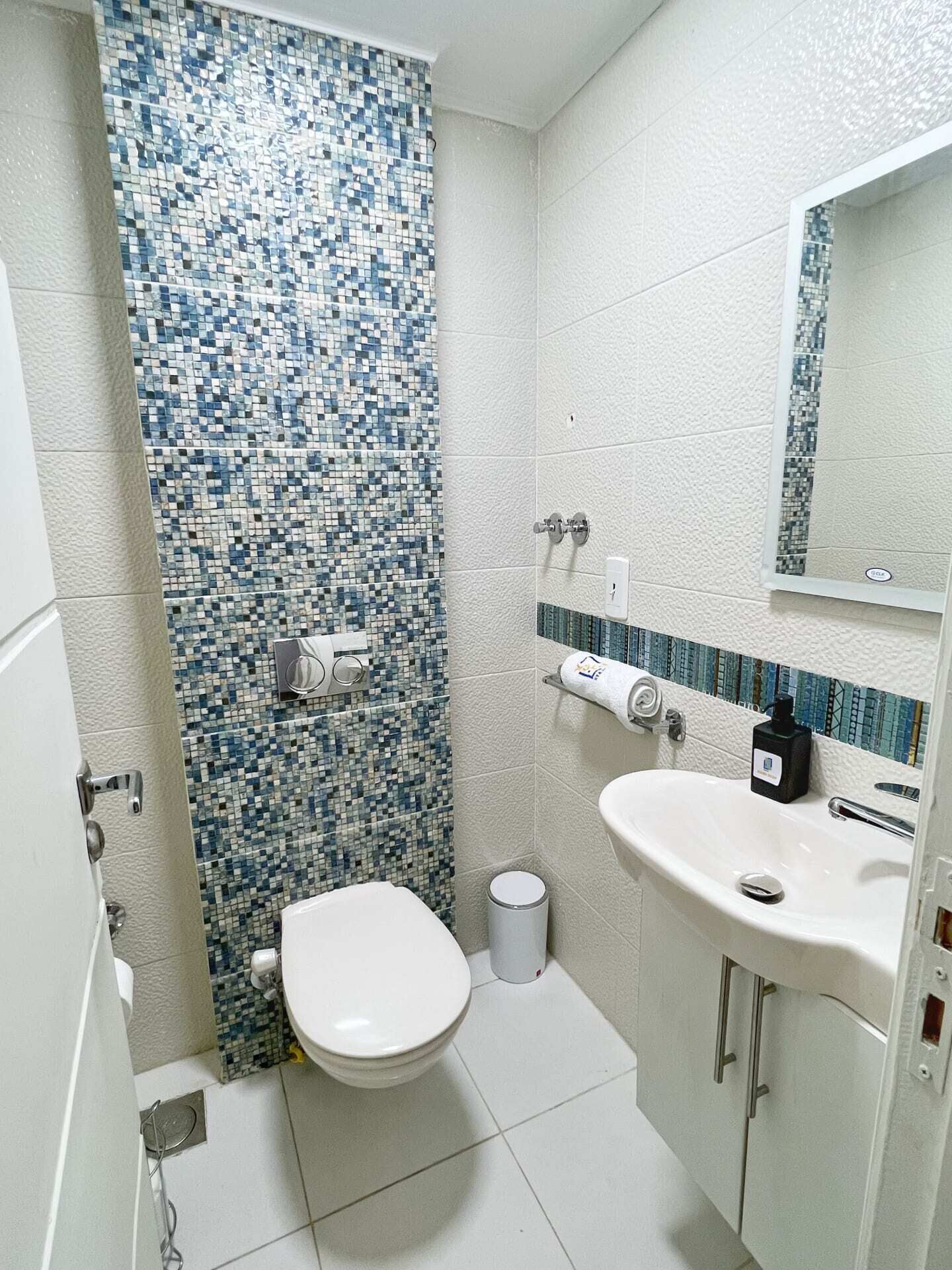 A bathroom with a mosaic tile shower wall, a toilet, a sink with a mirror, and a trash can.