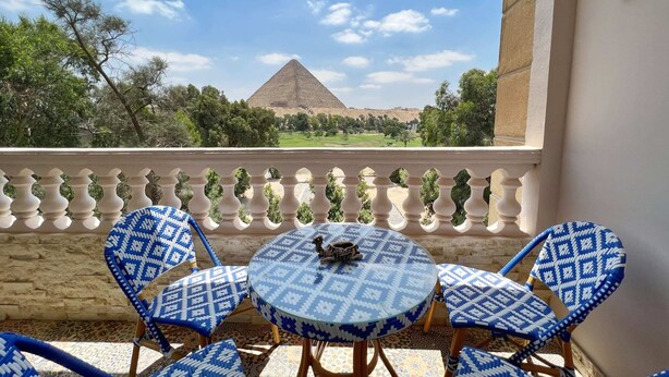 A balcony with a view of a pyramid, blue patterned chairs, and a small table with a decorative item. - Xuru Stays I Spellbinding 2 BDR Apt. At The Giza Pyramid's Doorstep (Giza)