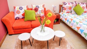 Ein gemütliches Zimmer mit einem roten Sofa, einem weißen runden Tisch mit zwei Stühlen und einem Bett mit bunter Bettwäsche.
