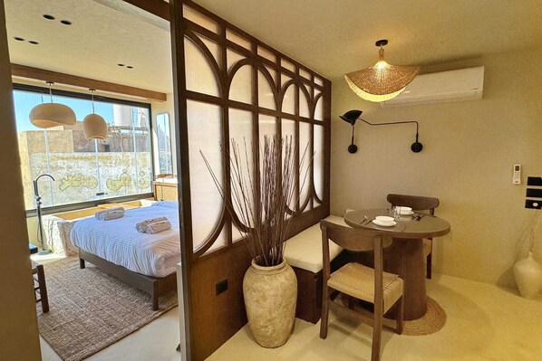A room with a bed, a dining table with chairs, a vase with branches, a wall-mounted lamp, and a window with a view of a building. - Xuru Stays I The Pyramids Horizon Studio w/ Bathtub (Giza)