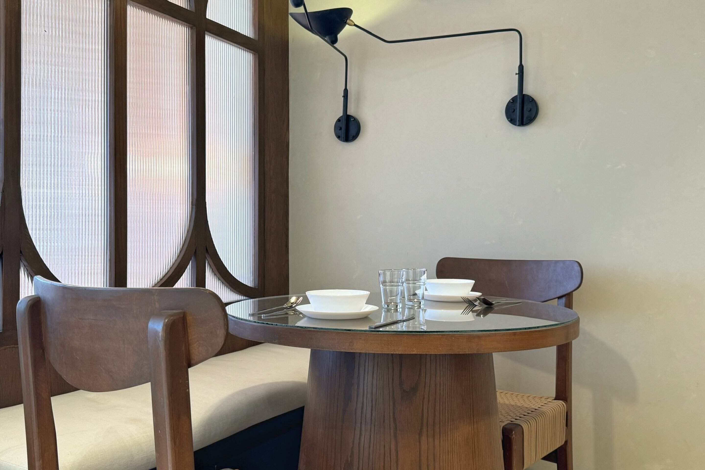 A round glass table with a wooden base, accompanied by two wooden chairs with woven seats. The table is set with white plates, bowls, and glasses.