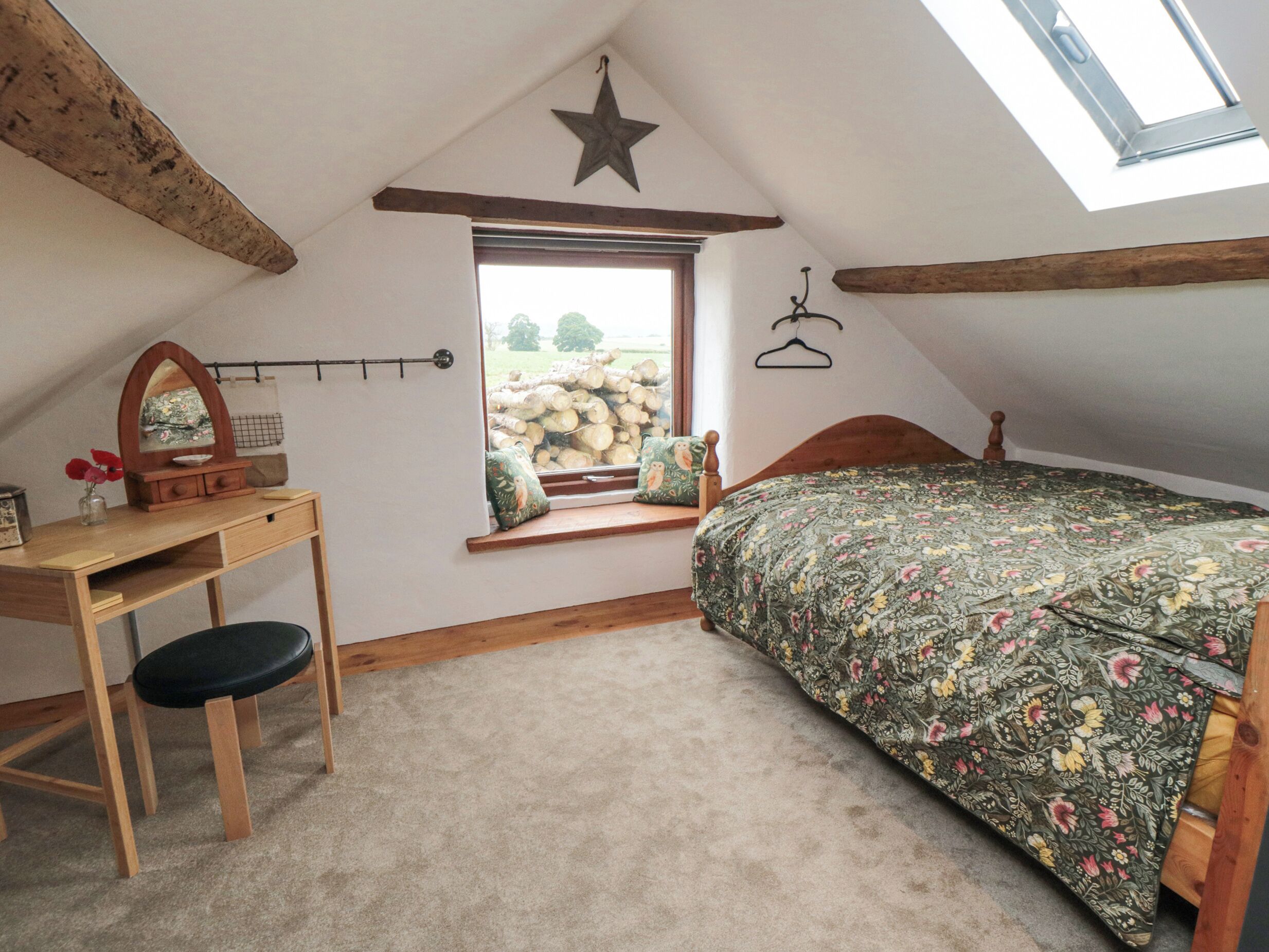 A cozy attic bedroom with a wooden desk, a bed with floral bedding, a star decoration, and a window with a view of trees and logs.