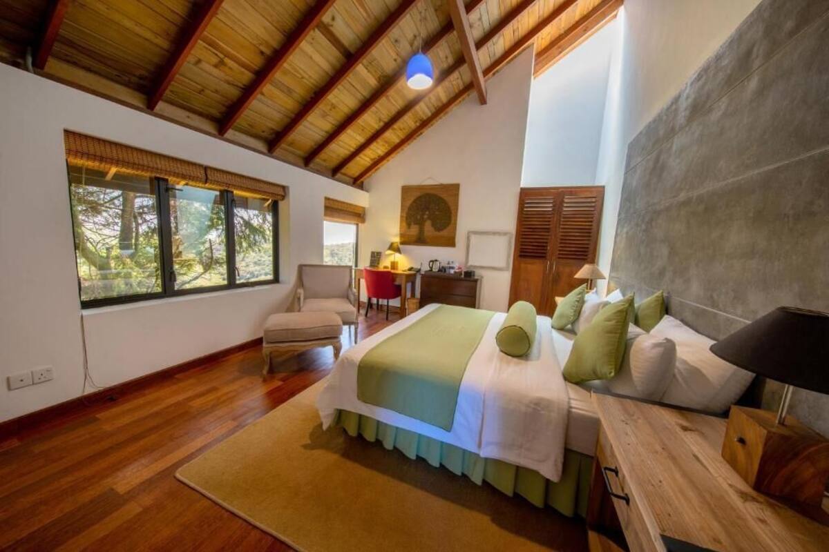 A bedroom with a large bed, a desk, and a chair. There are windows with a view of trees.
