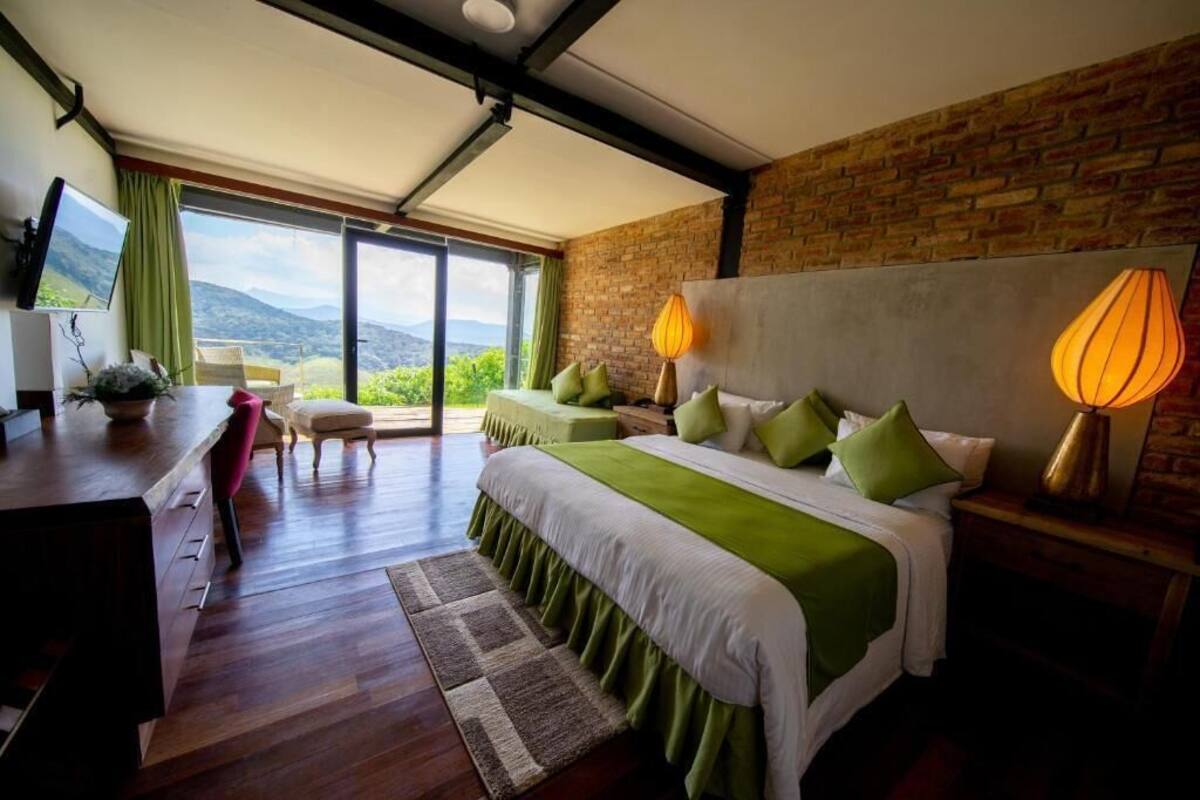 A bedroom with a large bed, green bedding, two bedside lamps, a desk with a computer, and a view of the outdoors.