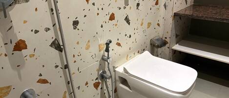A bathroom with a white toilet, a shower with a metal hose, and a wall with a terrazzo finish.