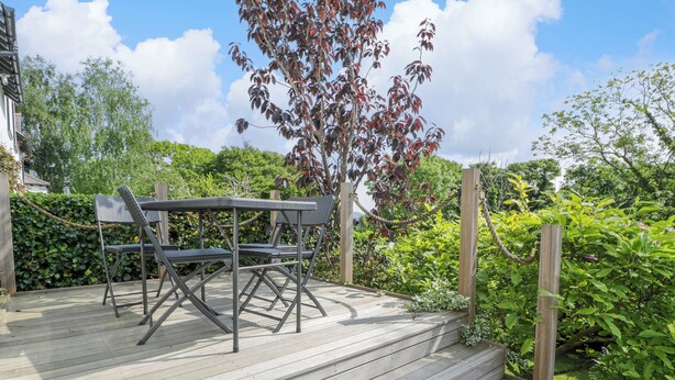 A wooden deck with a table and chairs, surrounded by lush greenery and a tree with red leaves. - Bron Y Graig (Conwy)