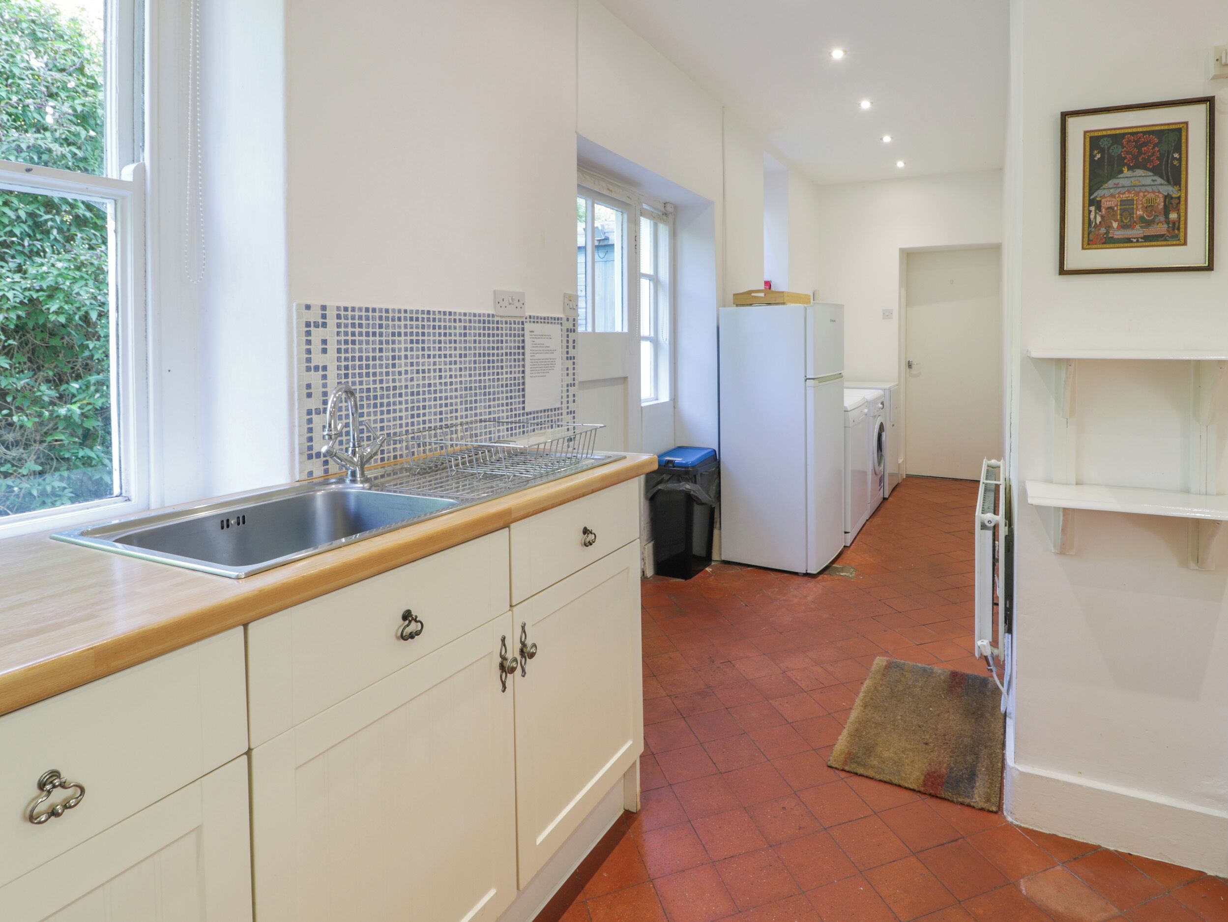 Een keuken met witte kastjes, een roestvrijstalen spoelbak, een betegelde spatwand, een wasmachine en een koelkast.