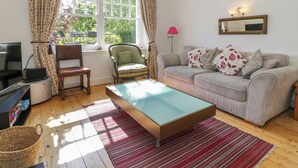 A living room with a sofa, armchair, and a glass-top coffee table. There is a television on a stand, a lamp, and a window with curtains. - Bron Y Graig (Conwy)