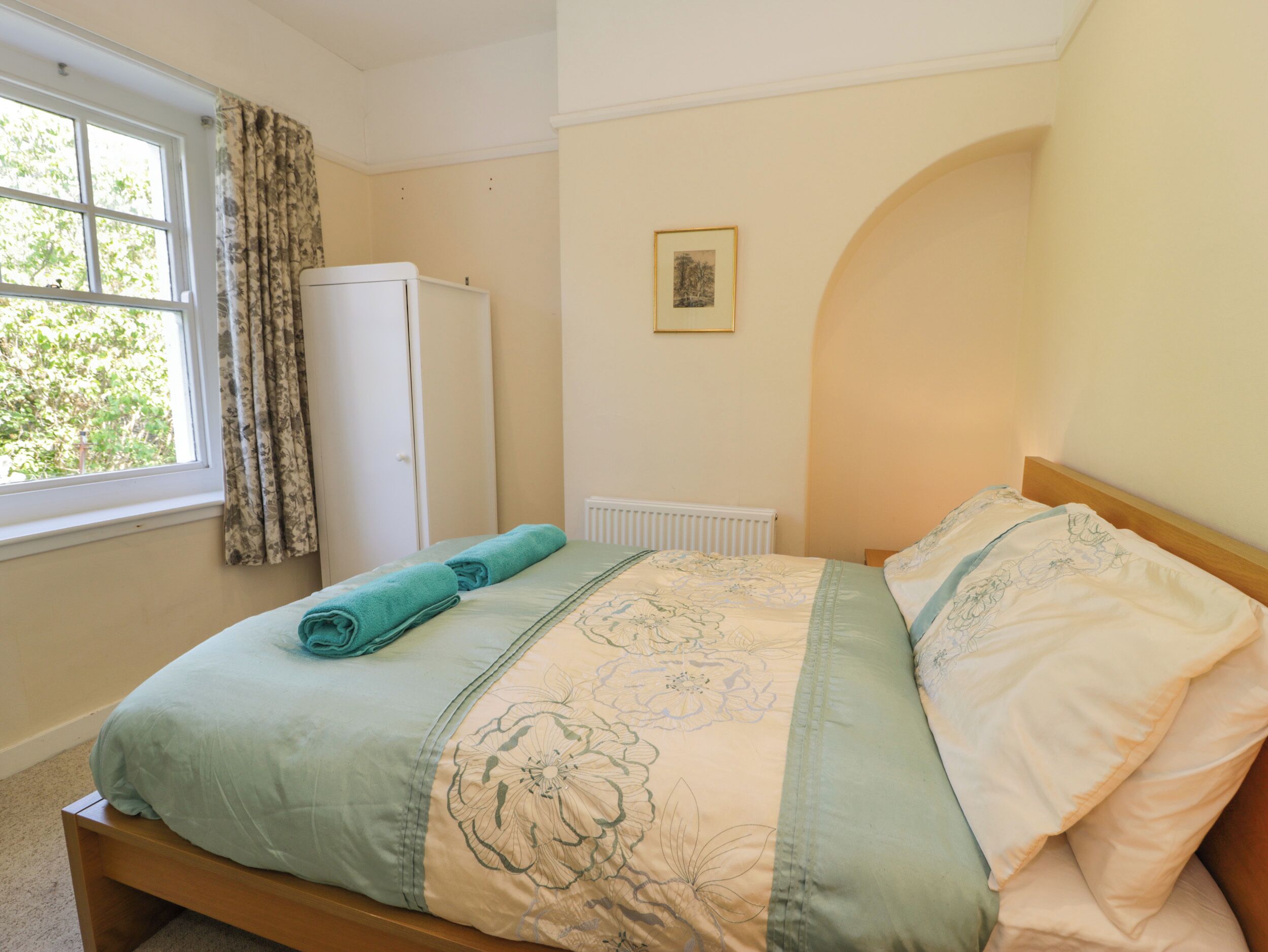 A neatly made bed with floral bedding, a window with curtains, a radiator, and a framed picture on the wall.