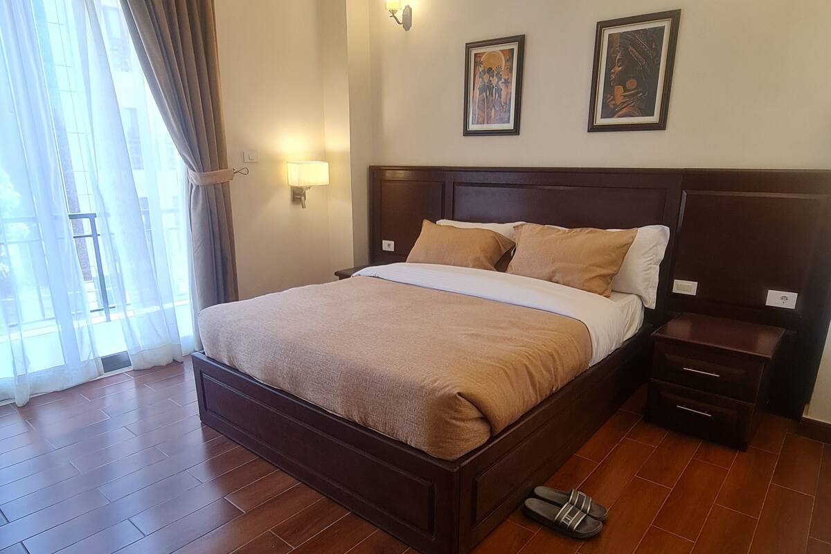 A hotel room with a large bed, wooden flooring, two bedside tables, a nightstand, a window with curtains, and wall-mounted lighting.