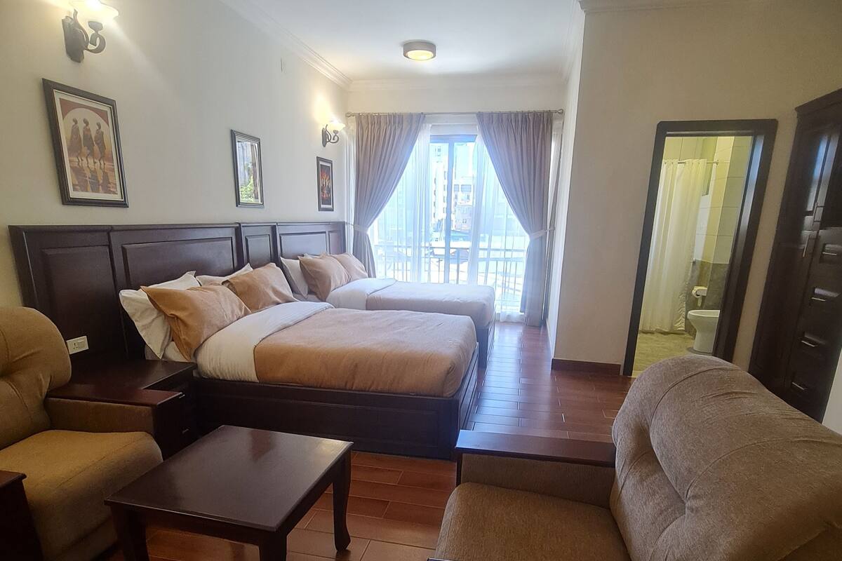 A hotel room with two beds, a sofa, a coffee table, and a view of the outside.