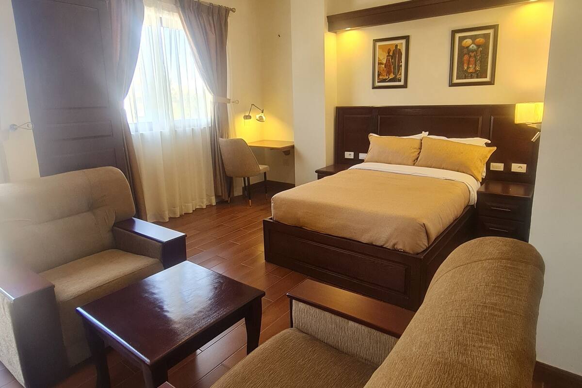 A hotel room with a bed, a sofa, a chair, a small table, and a nightstand with a lamp.
