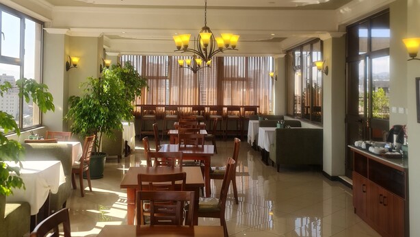 A spacious dining area with multiple tables and chairs, a large chandelier, and large windows allowing natural light. - FBK Guest House  (Addis Ababa)