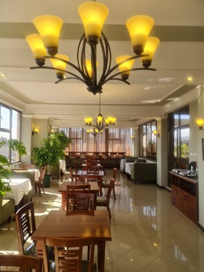 Uma sala de jantar ampla, com várias mesas e cadeiras, um lustre grande e janelas amplas que permitem a entrada de luz natural.