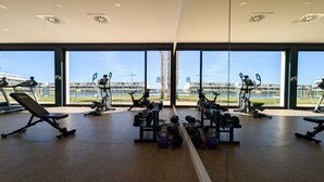 A gym with cardio equipment, weight machines, and dumbbells. It has large windows with a view of a building and a body of water. - CAMPORROSSO relax apartment (Finestrat)
