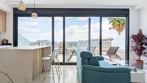 A modern living space with a kitchen area, a sofa, a dining table with chairs, and a large sliding glass door leading to an outdoor terrace with a view of the city. - CAMPORROSSO relax apartment (Finestrat)