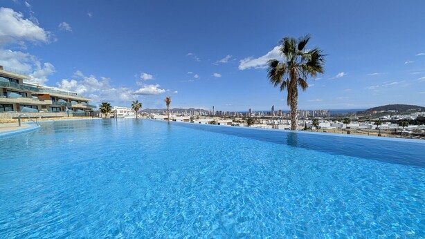 A clear blue infinity pool with a cityscape view, palm trees, and modern buildings in the background. - CAMPORROSSO relax apartment (Finestrat)