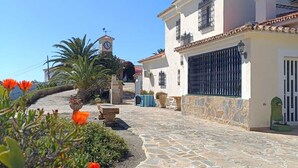 A stone-paved courtyard with a white building featuring a balcony and a clock tower in the background.