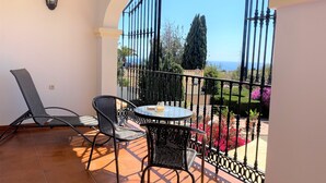 A balcony with a round table and two chairs, a wall-mounted light, and a view of a garden and distant landscape.