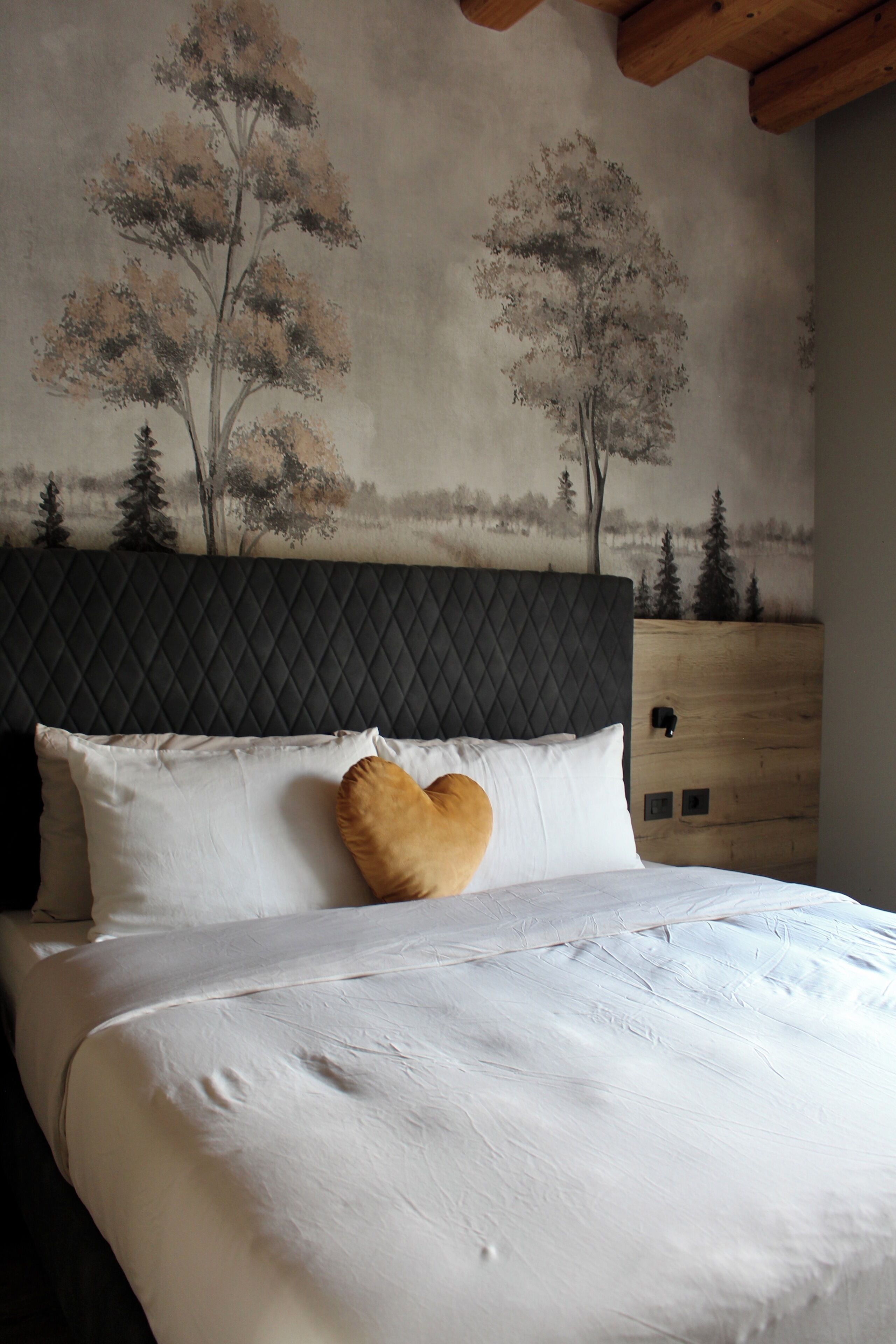 Una camera da letto con una testiera trapuntata, un letto con lenzuola bianche, un cuscino a forma di cuore e un affresco raffigurante alberi.