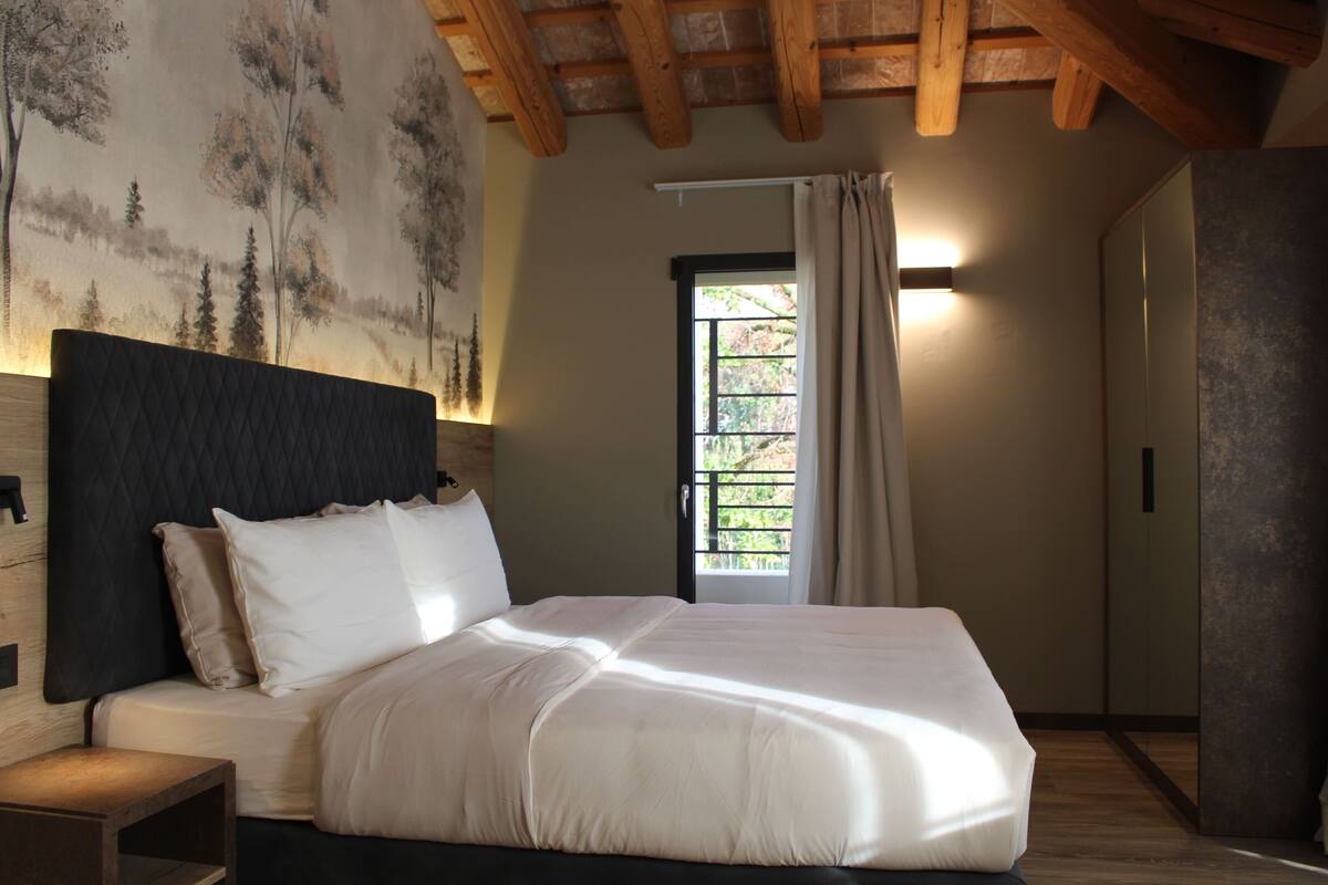 A bedroom with a large bed, a headboard, a nightstand, a window with curtains, and a wall with a nature-themed mural.