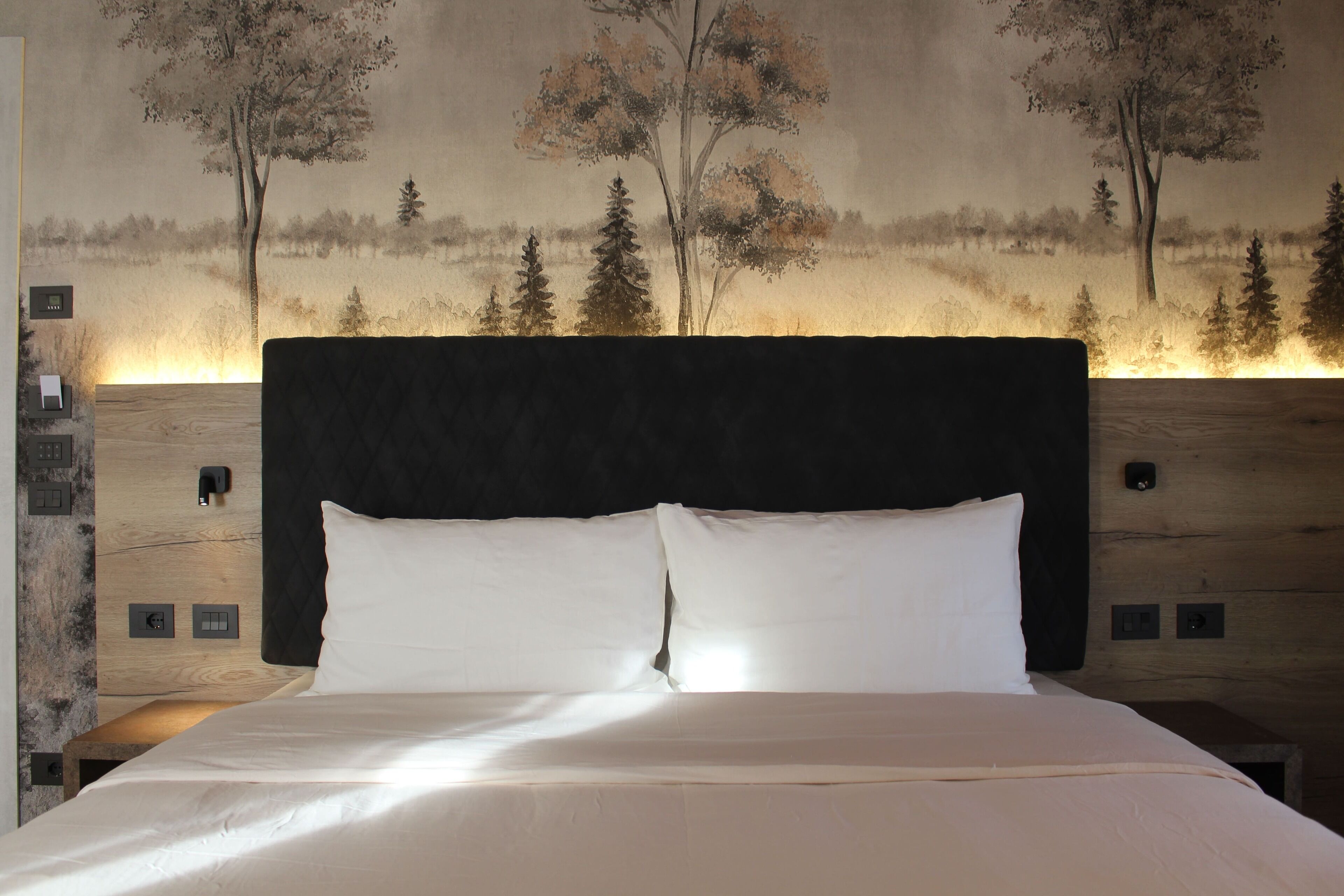 A bed with white linens, a dark headboard, and a wall mural of trees.